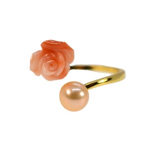 May include: A gold-toned open ring featuring a carved peach-colored coral rose on one end and a smooth, round, peach-colored pearl on the other end. The band is thin and curved, connecting the two distinct decorative elements in a bypass style.