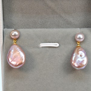 Puede incluir: Un par de pendientes colgantes con una gran perla rosa irregular e iridiscente en forma de gota y una perla rosa más pequeña en la parte superior. Los pendientes tienen postes y cadenas de color dorado.