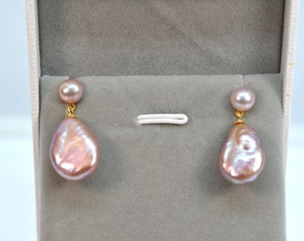Può includere: Un paio di orecchini pendenti con un grande ciondolo a goccia di perla rosa irregolare e iridescente e una perla rosa più piccola in cima. Gli orecchini hanno perni e catene dorati.