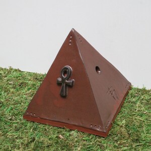 May include: A brown ceramic pyramid sculpture featuring a raised ankh symbol on the front face, a small circular hole near the top, and decorative dot patterns at the base corners. The surface has a smooth, glazed finish with subtle color variations.