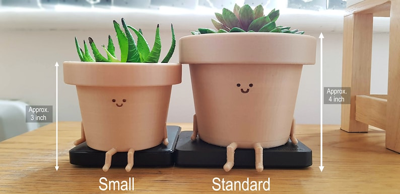 May include: Two terracotta-colored ceramic planters with smiling faces and legs, sitting on black bases. The smaller planter is approximately 3 inches tall and the larger planter is approximately 4 inches tall.  The planters are ideal for small plants.