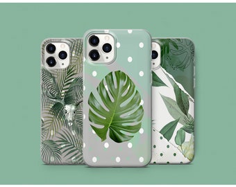 May include: Three clear phone cases with different tropical leaf designs. The first case has a white skull and palm leaves on a gray background. The second case has a large green monstera leaf on a gray background with white polka dots. The third case has a white background with green leaves and a ripped paper effect.