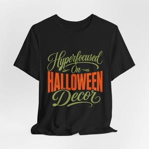 May include: A black short-sleeve t-shirt featuring the text Hyperfocused On HALLOWEEN Decor. The word Hyperfocused and Decor are written in a light green cursive script, while the word HALLOWEEN is printed in large, bold, orange block letters.