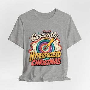 Op de afbeelding: Een grijs gemêleerd t-shirt met een afbeelding van een kleurrijk kerstornament in de vorm van een doelwit met een gele bliksemschicht. De tekst luidt Currently HYPERFOCUSED ON CHRISTMAS in een vet, meerkleurig retro lettertype.