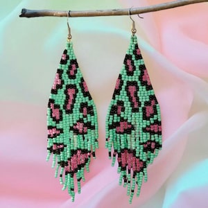 May include: A pair of beaded earrings with a leopard print design. The earrings have a triangular shape with a mint green background, black spots, and pink accents. The fringe at the bottom adds movement and flair.