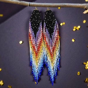 May include: A pair of beaded earrings with a black top section and a colorful, fringed bottom. The fringe transitions from blue to yellow, orange, and purple. The earrings are hanging from a small branch against a dark background.