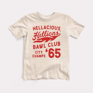 May include: A beige t-shirt with a red graphic that says "Hellacious Hellions Bawl Club City Champs '65". The graphic features a flame design.
