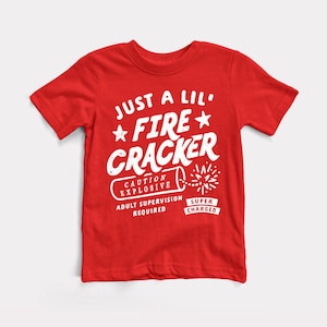 May include: A red t-shirt with white text that reads "JUST A LIL' FIRECRACKER" with a firecracker graphic. The shirt also includes the phrases "CAUTION EXPLOSIVE", "ADULT SUPERVISION REQUIRED", and "SUPER CHARGED".