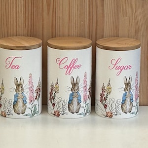 以下が含まれることがあります： 木製の蓋が付いた白いセラミックキャニスター3つ。「Tea」、「Coffee」、「Sugar」とピンクの筆記体でラベル付けされています。各キャニスターには、青いジャケットを着たウサギが花の中にいる水彩画のイラストが描かれています。