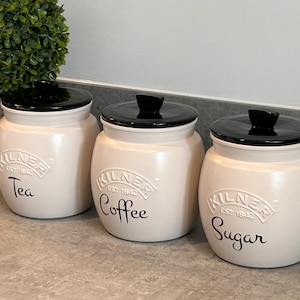 May include: Three white ceramic canisters with black lids and black lettering. The canisters are labelled "Tea", "Coffee", and "Sugar".