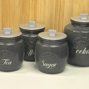 May include: Four matching gray ceramic kitchen canisters with silver lids. The canisters are labeled "Tea", "Coffee", "Sugar", and "Cookies" in a decorative font. The canisters are arranged in a row against a light wood background.