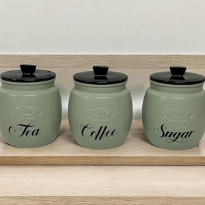 Può includere: Tre barattoli di ceramica verde con coperchi neri e scritte nere. I barattoli sono etichettati con "Tè", "Caffè" e "Zucchero".