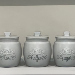 May include: Three grey ceramic canisters with white lids and black lettering. The canisters are labelled "Tea", "Coffee", and "Sugar".