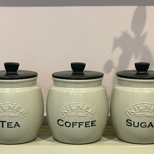 May include: Three ceramic canisters with black lids and green bodies. The canisters are labeled "Tea", "Coffee", and "Sugar" with the Kilner logo and "Est. 1842".