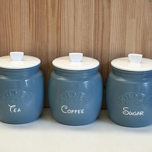May include: Three matching blue ceramic canisters with white lids. The canisters are labeled "Tea", "Coffee", and "Sugar" in white script. The brand name "Kilner" is embossed on each canister.