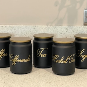 May include: Five black canisters with gold lids and gold lettering. The canisters are labelled "Coffee", "Coffee Mate", "Tea", "Herbal Tea", and "Sugar".