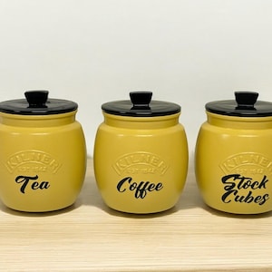 Pode incluir: Três potes de cerâmica amarelos com tampas pretas e letras pretas. Os potes são etiquetados com "Chá", "Café" e "Cubos de Caldo".