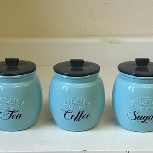 Puede incluir: Tres botes de cerámica azul claro con tapas negras. Cada bote tiene una etiqueta negra con las palabras "Tea", "Coffee" y "Sugar" en escritura cursiva. Los botes son de la marca Kilner.