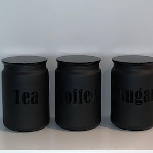 May include: Three black kitchen storage jars with lids. Each jar is labelled with a word: "Tea", "Coffee", and "Sugar". The jars are cylindrical and sit on a white shelf against a white wall. The text is in a simple font.