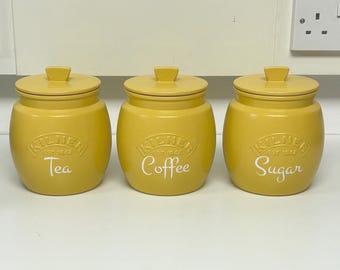 May include: Three yellow ceramic kitchen canisters with lids, labeled "Tea," "Coffee," and "Sugar" in white script. Each canister has a rounded shape and a square knob on the lid. The brand name "Kilner" is embossed on each.