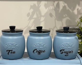 Op de afbeelding: Drie blauwe keramische potten met zwarte deksels en zwarte letters. De potten zijn gelabeld met "Thee", "Koffie" en "Suiker".