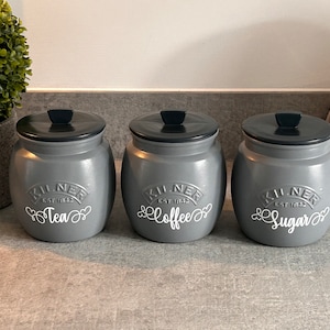 Puede incluir: Tres botes grises Kilner con tapas negras, etiquetados "Tea", "Coffee" y "Sugar" en escritura blanca. Los botes están colocados sobre una encimera, con una planta y un electrodoméstico blanco al fondo.