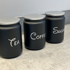 Op de afbeelding: Drie zwarte keukenpotten met zilveren deksels. De potten zijn voorzien van de tekst "Tea", "Coffee" en "Sugar" in witte letters. De potten zijn cilindrisch en staan op een grijs aanrecht.