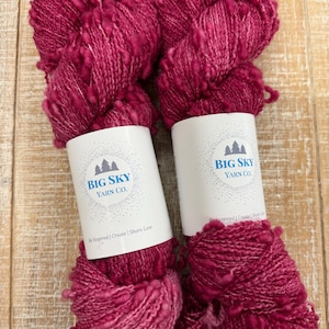 May include: Two skeins of vibrant magenta yarn from Big Sky Yarn Co. are displayed. The yarn has a textured, looped appearance. The white labels feature a mountain logo and the text "Be Inspired | Create | Share Love".