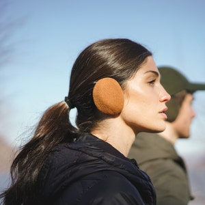 May include: A woman with long dark hair wearing a black jacket with a brown ear warmer on her left ear.