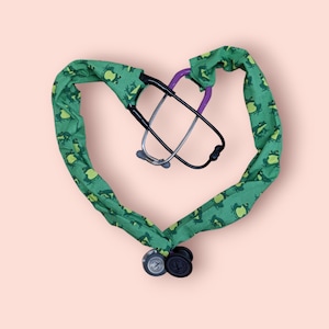 May include: A green stethoscope cover with a frog pattern. The cover is shaped like a heart and has a purple stethoscope inside.