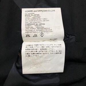May include: A close-up of a tag from a Comme des Gar&ccedil;ons garment. The tag is white with black text, listing fabric composition, care instructions, and the text "MADE IN JAPAN". The tag also includes Japanese text and care symbols.