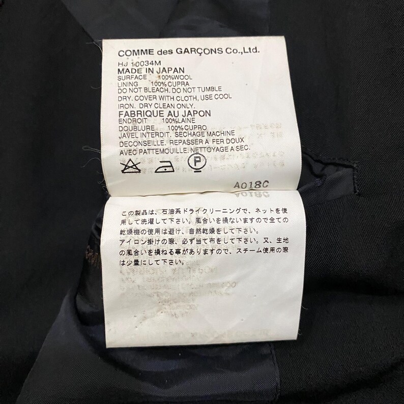 May include: A close-up of a tag from a Comme des Gar&ccedil;ons garment. The tag is white with black text, listing fabric composition, care instructions, and the text "MADE IN JAPAN". The tag also includes Japanese text and care symbols.
