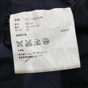 May include: A white clothing tag with Japanese text and symbols. The tag includes the product code "HJ 10034M", fabric composition details, size "M", and care instructions. The tag also features the text "AD2000".