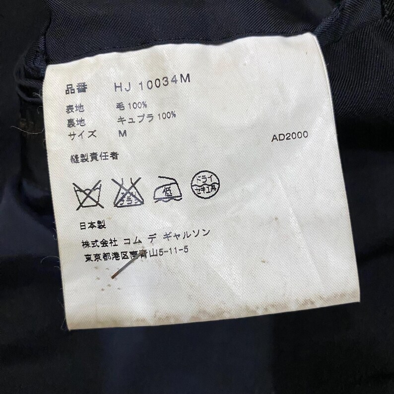 May include: A white clothing tag with Japanese text and symbols. The tag includes the product code "HJ 10034M", fabric composition details, size "M", and care instructions. The tag also features the text "AD2000".