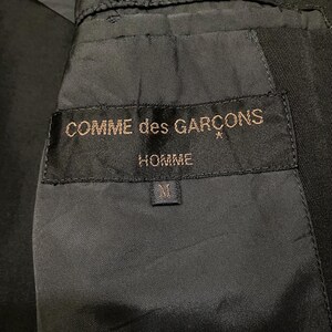 May include: Close-up of a black garment's interior, showcasing a label with the text "COMME des GARCONS HOMME" in gold lettering. The label also features a size "M" tag. The fabric appears to be a dark, possibly wool, material.