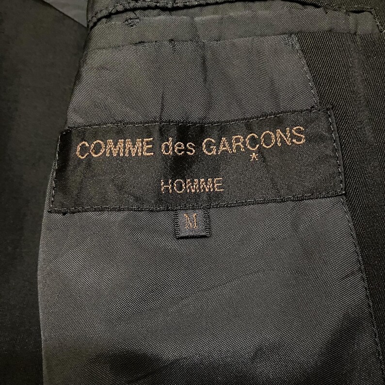 May include: Close-up of a black garment's interior, showcasing a label with the text "COMME des GARCONS HOMME" in gold lettering. The label also features a size "M" tag. The fabric appears to be a dark, possibly wool, material.