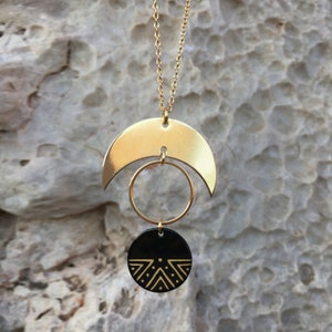 Puede incluir: Un collar dorado con una media luna, un c&iacute;rculo y un colgante circular negro. El colgante negro presenta un dise&ntilde;o geom&eacute;trico dorado. El collar cuelga sobre un fondo texturizado de color claro.