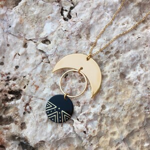 Puede incluir: Collar dorado con un colgante de media luna, un elemento circular y un disco negro con un dise&ntilde;o geom&eacute;trico blanco. La cadena es delicada, creando un accesorio de tem&aacute;tica celestial.