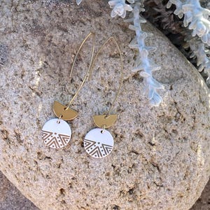 Può includere: Un paio di orecchini color oro dal design moderno. Ogni orecchino presenta un semicerchio dorato e un pendente circolare bianco con un motivo geometrico in un colore oro più scuro. Gli orecchini sono esposti su una superficie di pietra strutturata.