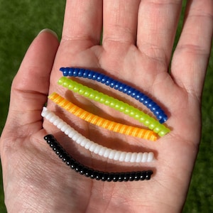 Może przedstawiać: Dłoń trzyma pięć kolorowych, zwiniętych plastikowych sznurk&oacute;w. Sznurki są niebieskie, zielone, pomarańczowe, białe i czarne. Sznurki wykonane są z małych, okrągłych koralik&oacute;w nanizanych razem. Tło to rozmyty zielony trawnik.