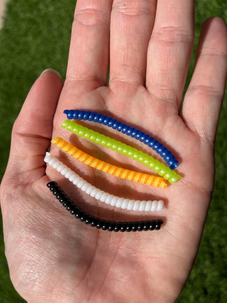 Może przedstawiać: Dłoń trzyma pięć kolorowych, zwiniętych plastikowych sznurk&oacute;w. Sznurki są niebieskie, zielone, pomarańczowe, białe i czarne. Sznurki wykonane są z małych, okrągłych koralik&oacute;w nanizanych razem. Tło to rozmyty zielony trawnik.