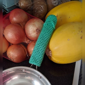 May include: A green crocheted pot holder on a countertop next to a yellow squash and a bag of red onions.