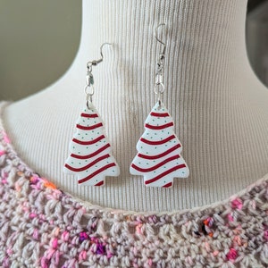 May include: A pair of white Christmas tree earrings with red and green stripes and white sprinkles. The earrings are hanging from silver hooks.