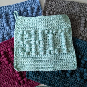 May include: Five crocheted pot holders in different colors. The pot holders have a textured, bumpy surface. One pot holder is light blue, one is gray, one is teal, one is burgundy, and one is light green. The light green pot holder has the word "YOU" crocheted into it.