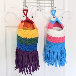 May include: Two handmade, whimsical, crocheted storage bags. One is rainbow-colored with purple fringe, the other features pink, white, and blue stripes with blue fringe. Both have large googly eyes and are hanging from a metal rack.