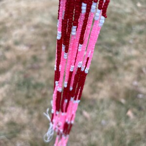 May include: Close-up of strands of beads in shades of red, pink, and white. The beads are small and strung together, creating a textured effect. The background is blurred, suggesting an outdoor setting. The beads are likely used for jewelry or craft projects.