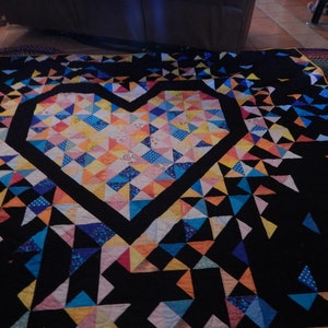 May include: A black quilt with a large heart-shaped design in the center. The heart is made of colorful patchwork fabric with a variety of patterns and colors, including yellow, blue, pink, and orange. The quilt is made of many small triangles.
