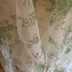 May include: A sheer, lightweight fabric garment featuring a delicate botanical print in shades of green on a white background. The material is draped in soft folds, showcasing a repeating pattern of various leaves, stems, and small flowers.