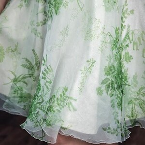 May include: A knee-length skirt made of a sheer, off-white fabric featuring a printed pattern of green botanical elements, including various leaves, stems, and small flowers. The skirt has a flared, flowing silhouette with a layered, translucent appearance.
