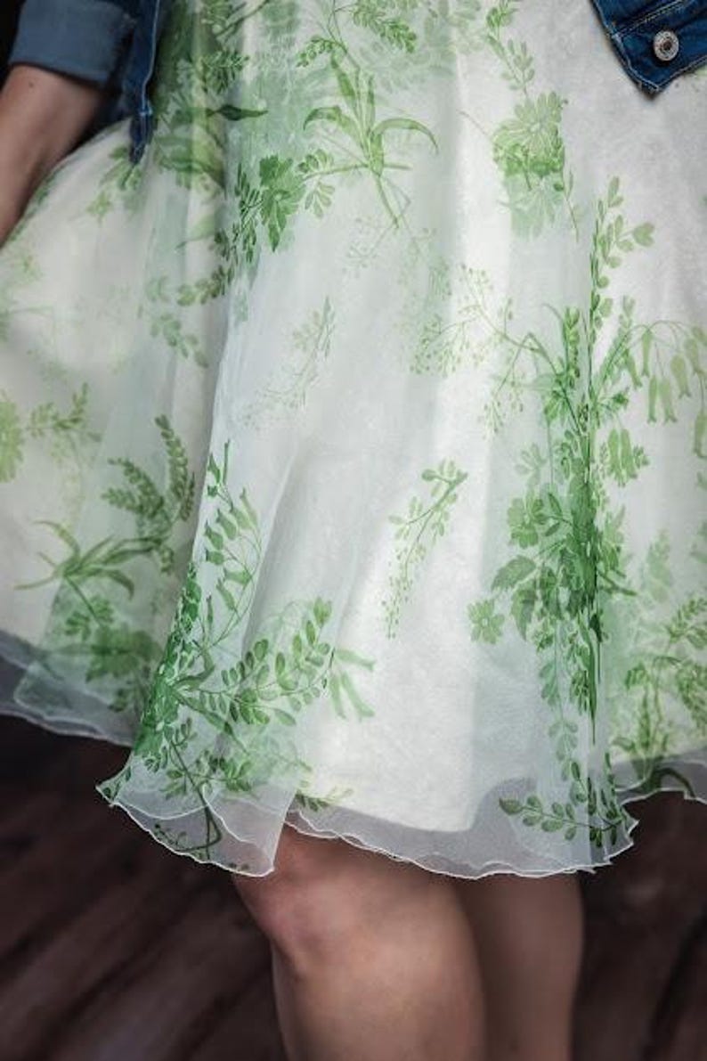 May include: A knee-length skirt made of a sheer, off-white fabric featuring a printed pattern of green botanical elements, including various leaves, stems, and small flowers. The skirt has a flared, flowing silhouette with a layered, translucent appearance.